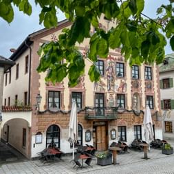 Traditional Bavarian building with colorful facade paintings in Garmisch. Outdoor seating with umbrellas in front, framed by green tree leaves.