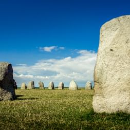 Antikes Steinmonument Ales Stenar mit großen Steinen in ovaler Formation auf Grasfeld unter blauem Himmel mit weißen Wolken.