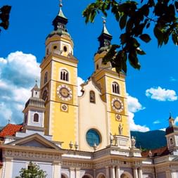 Brixen Cathedral with yellow and white baroque facade, twin bell towers with clocks, framed by green leaves against a blue sky with clouds.