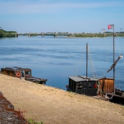 Traditionelle Holzboote liegen an einer Steinmauer an der Loire vertäut. Eine Brücke überspannt das ruhige blaue Wasser im Hintergrund.