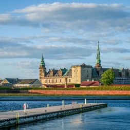 Kronborg Castle in Helsingør with towers and copper roofs behind red fortress walls. A wooden pier extends into the blue water in the foreground.