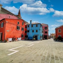 Sonniger Platz in Pellestrina mit bunten Häusern in Orange, Blau, Rosa und Rot. Gepflasterter Platz mit Kirchturm und blauem Himmel mit Wolken.