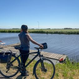 Radfahrerin mit Fahrrad steht auf Holzsteg am Kanal und blickt auf flache niederländische Landschaft mit grünen Feldern unter blauem Himmel.