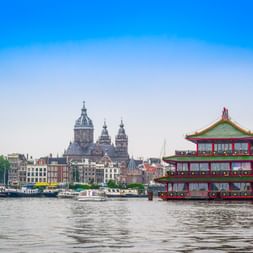 Amsterdamer Hafenansicht mit schwimmendem Restaurant im chinesischen Stil und St. Nikolaus Kirche im Hintergrund. Historische Gebäude säumen das Ufer.
