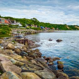 Rocky shoreline with colorful houses on Ven island. Calm blue water, wooden pier, and green hills under cloudy sky.