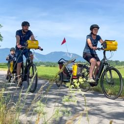 Family of three cycling on paved path with yellow panniers. Mountains and green meadows visible in background under blue sky.