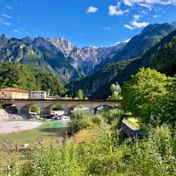 Stone bridge crossing a turquoise river in Kanaltal valley, surrounded by lush green vegetation and dramatic Alpine mountain peaks under blue sky.
