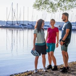 Three cyclists with helmets standing at the shore of Starnberger See. Marina with sailboats and mountains visible in background.
