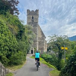 Radfahrer in türkisfarbenem Shirt fährt auf eine Steinkirche mit Turm in Sankt Michael, Wachau zu. Gelbe Wegweiser entlang des Weges.