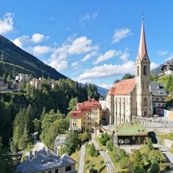 Church with red spire in Bad Gastein surrounded by colorful buildings on hillside. Alpine mountains and forests frame the Austrian spa town.