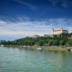 Burg Bratislava auf einem grünen Hügel mit Blick auf die Donau. Schiffe liegen am Flussufer unter blauem Himmel mit weißen Wolken.