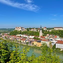 Panoramic view of Burghausen with its historic castle complex on a hilltop, colorful town buildings, and a green river in the foreground.