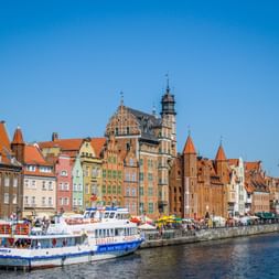 Colorful historic buildings along Gdansk waterfront with tour boats moored at the promenade. Medieval crane tower visible on the right under clear blue sky.