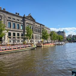Grachtenboote am Rokin in Amsterdam mit historischen Gebäuden und Bäumen entlang des Wassers unter blauem Himmel.