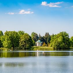 Blick über einen ruhigen See mit Spiegelung von Bäumen und Himmel, mit einem kleinen türkisfarbenen Pavillon umgeben von grünen Bäumen in Kassel.