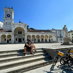 Two cyclists sitting on steps in Piazza della Libertà, Udine, with bicycles and yellow panniers nearby. Historic clock tower and arcaded buildings visible.