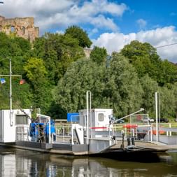 Autofähre auf der Weser mit der Burgruine Polle auf einem Hügel im Hintergrund, umgeben von grünen Bäumen unter blauem Himmel mit Wolken.