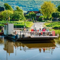 Cyclists with bikes on a ferry crossing the Weser River at Hemeln. Village with half-timbered houses and green hills visible in background.