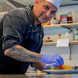 Smiling chef in white hat and blue gloves plating food in a ship kitchen. Shelves with ingredients visible in background.