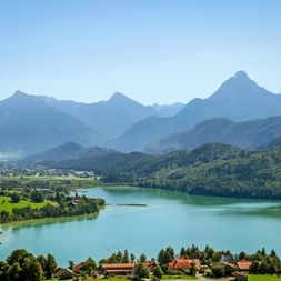 Weissensee mit türkisfarbenem Wasser, umgeben von grünen Wiesen und Wäldern, mit Allgäuer Alpengipfeln im Hintergrund unter klarem Himmel.