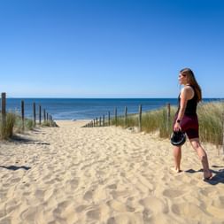 Woman in cycling attire holding helmet stands on sandy beach path with wooden fence posts, dune grass, and ocean view under clear blue sky.