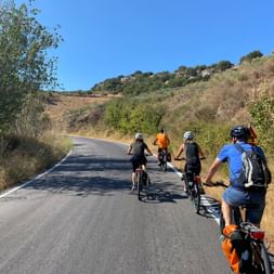 Gruppe von Radfahrern auf einer asphaltierten Straße durch hügeliges Gelände auf Kreta. Die Landschaft zeigt trockenes Gras und grüne Vegetation unter blauem Himmel.