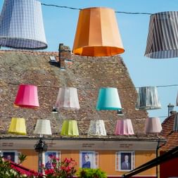 Colorful decorative lampshades hanging above a historic street in Szentendre. Traditional buildings with tiled roofs and portraits in windows.