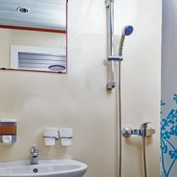Kompaktes Badezimmer auf dem Schiff MS Macek mit weißem Waschbecken, Spiegel, Dusche mit Vorhang und Chromarmaturen an cremefarbenen Wänden.