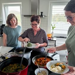 Three women cooking together in a modern kitchen, preparing colorful dishes in pans with vegetables and rice visible on the counter.
