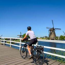 Radfahrerin auf Holzbrücke mit Blick auf traditionelle holländische Windmühlen in Kinderdijk entlang eines Kanals unter blauem Himmel.