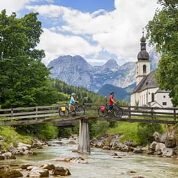 Two cyclists crossing a wooden bridge over a mountain stream in Ramsau, Berchtesgaden. White church with onion dome and Alpine peaks in background.
