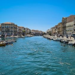 Canal lined with historic buildings and fishing boats in Sète. Blue water reflects clear sky, people walk along waterfront promenades.