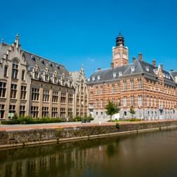 Historic buildings along the canal in Dendermonde, featuring ornate architecture with a prominent red-brick town hall with tower under blue sky.