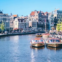Amsterdamer Gracht mit Ausflugsbooten und bunten historischen Gebäuden. Menschen entspannen auf der Uferpromenade unter blauem Himmel.