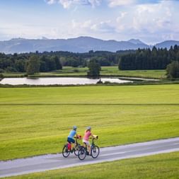 Two cyclists on a paved path through green meadows with a lake and mountains in the background in the Seenland region of Austria.