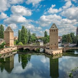 Medieval towers and bridges spanning a canal in Strasbourg. Two stone towers flank covered bridges over calm water reflecting clouds and trees.