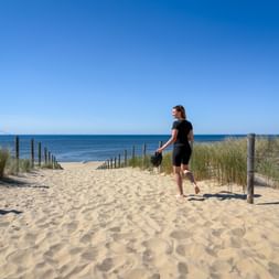 Radfahrerin in schwarzer Sportkleidung geht barfuß auf sandigem Strandweg in Zeeland, Helm in der Hand. Holzpfosten säumen den Weg zum Meer.