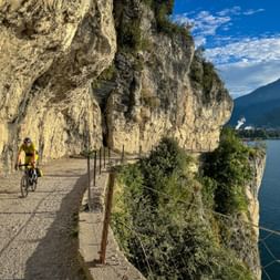 Radfahrer in gelbem Trikot auf der malerischen Strada del Ponale, die in die Klippe über dem Gardasee gehauen ist, mit Bergen und blauem Himmel.