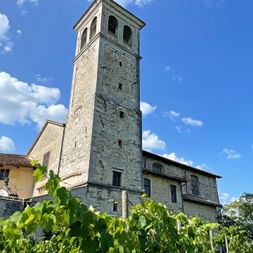 Historischer steinerner Glockenturm mit Bogenfenstern erhebt sich über einem Kirchengebäude in Cividale del Friuli, mit grünem Weinberg im Vordergrund.