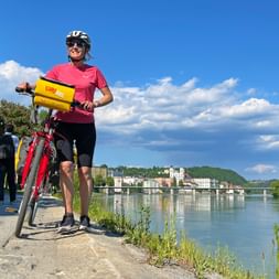 Radfahrerin in pinkem Trikot mit rotem Rad und gelber Tasche am Inn-Ufer in Passau, mit historischer Stadt und Kirche am gegenüberliegenden Ufer.