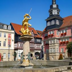Golden statue of St. George slaying dragon atop fountain in Eisenach market square, with colorful half-timbered houses and clock tower.