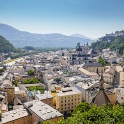 Panoramablick auf Salzburgs Altstadt mit historischen Gebäuden, Kirchenkuppeln, Festung Hohensalzburg auf dem Hügel und Alpenbergen im Hintergrund.
