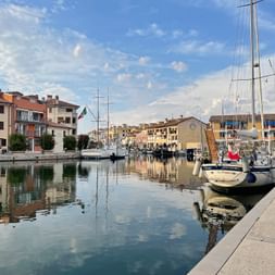 Peaceful harbor canal in Grado with moored sailboats, colorful buildings lining the waterfront, and reflections in the calm water under a blue sky.