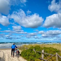 Cyclist in blue jacket with bicycle on sandy path leading to Baltic Sea beach. Wooden fence, dune grasses, waves and cloudy blue sky visible.