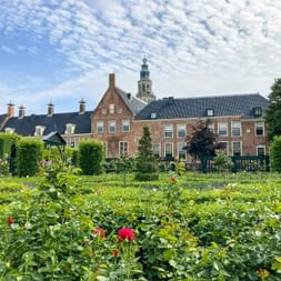 Prinsentuin-Garten in Groningen mit blühenden Pflanzen und historischen Backsteingebäuden. Ein Kirchturm ragt im Hintergrund unter bewölktem Himmel auf.
