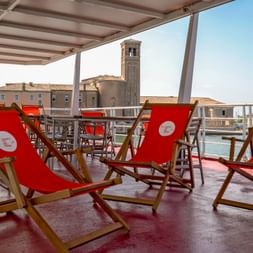 Sun deck of MS Ave Maria with red deck chairs under a canopy, overlooking a canal and historic church tower in Portegrandi.