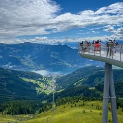 Moderne Aussichtsplattform auf der Schmittenhöhe mit Besuchern, die auf Alpental, See und Bergketten unter bewölktem Himmel blicken.
