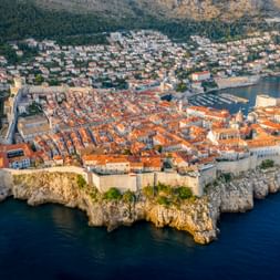 Aerial panorama of Dubrovnik's walled Old Town on a rocky peninsula, with terracotta roofs, historic fortifications, and blue Adriatic Sea.