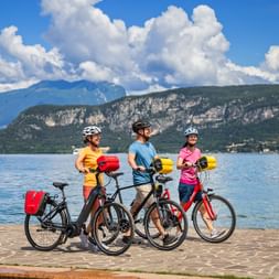 Three cyclists with helmets and panniers standing with bikes on Bardolino waterfront. Lake Garda and mountains visible under blue sky with clouds.