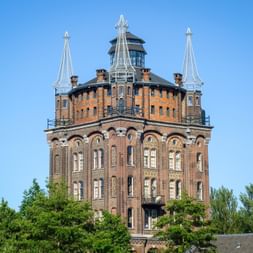 Historischer Backsteinwasserturm Villa Augustus in Dordrecht mit kunstvollen Verzierungen, drei weißen Türmchen und Bogenfenstern, umgeben von Bäumen.